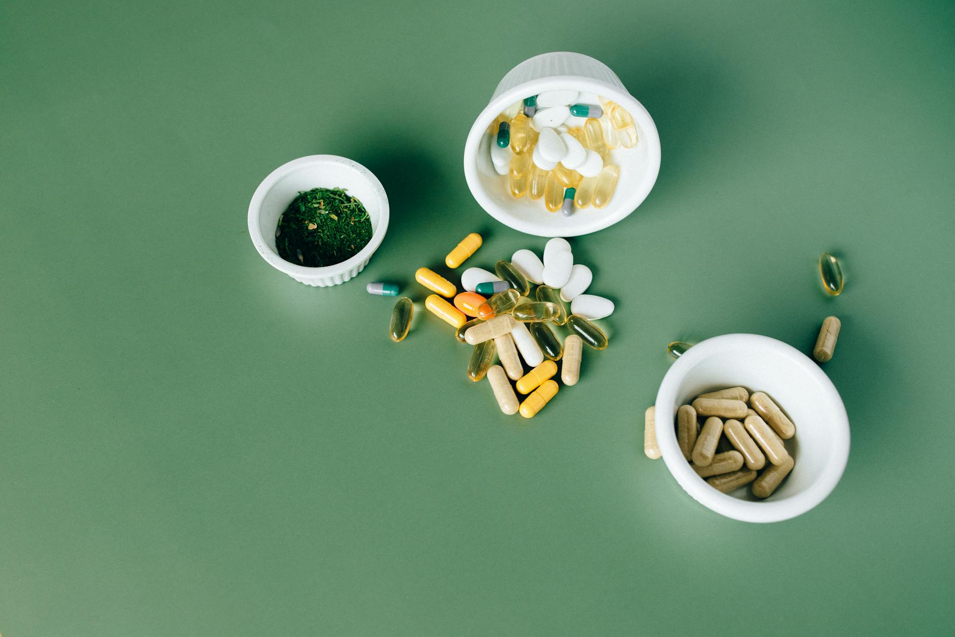Assorted vitamin capsules, tablets and softgels in white bowls on a green surface