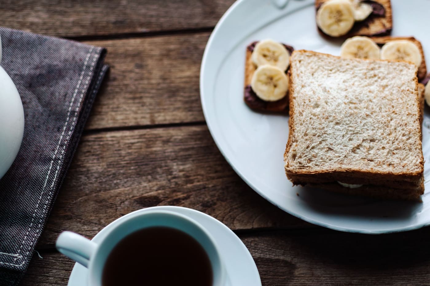 White toast and banana slices on a plate beside a cup of black coffee on a wooden table