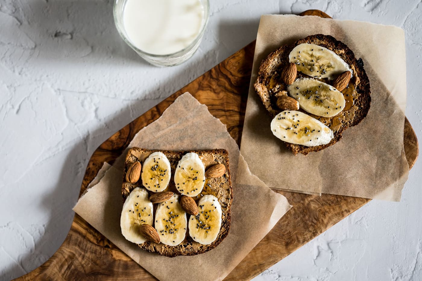 Two slices of toast topped with sliced banana, almonds and honey on a wooden board beside a glass of milk