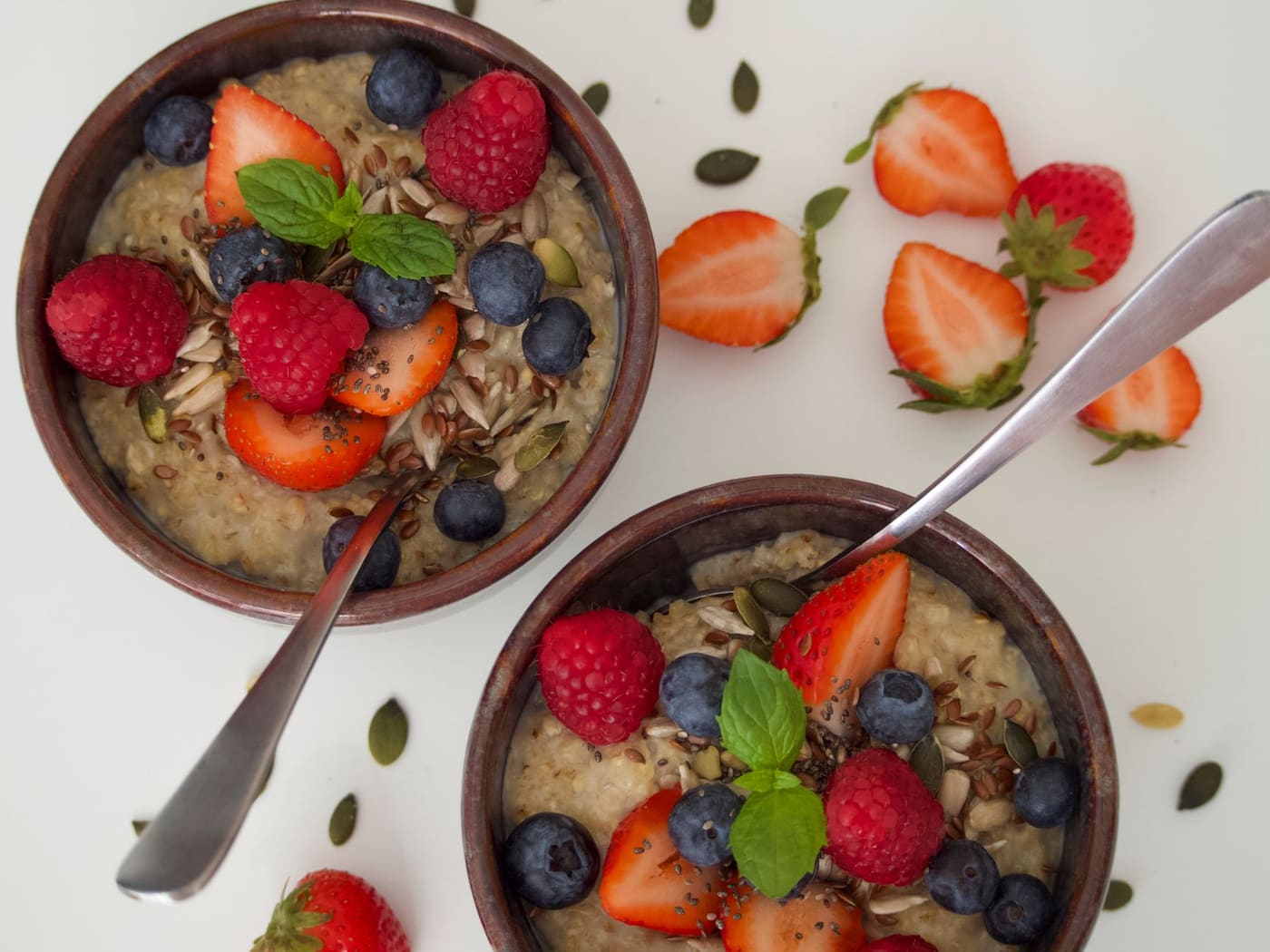 Two bowls of oatmeal topped with raspberries, blueberries, strawberries and seeds on a white surface
